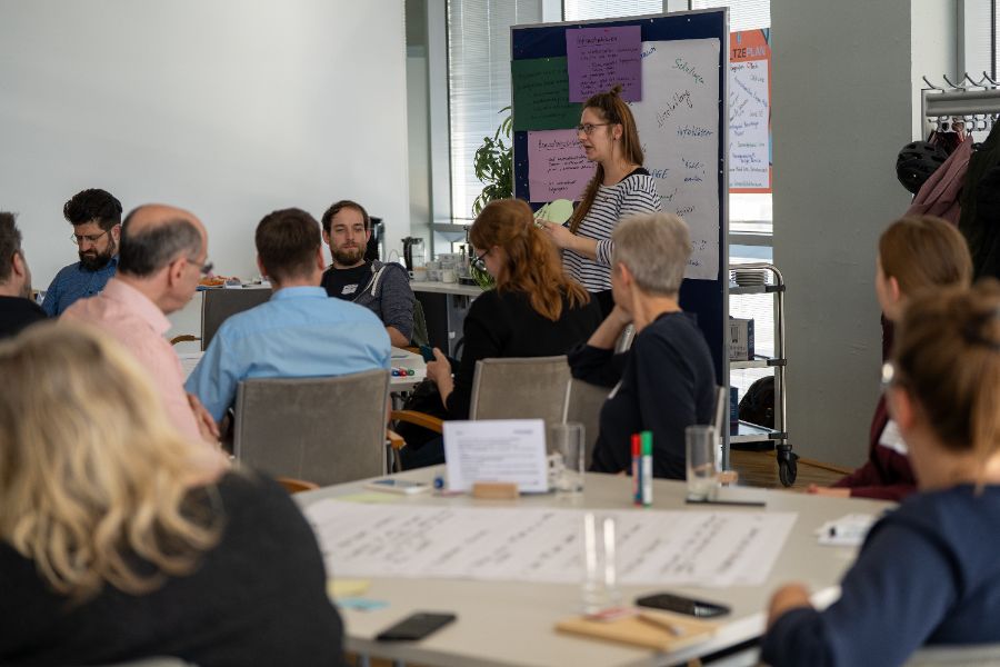 In einem Raum sitzende Personen und eine moderierende Person vor einer Pinnwand mit Plakaten, auf den Tischen liegen beschriebene Plakate und Stifte.
